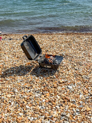 Unattended barbecue with flames from hot coals at the water's edge of a shale beach