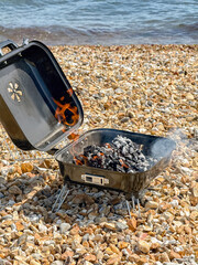 Unattended barbecue with flames from hot coals at the water's edge of a shale beach