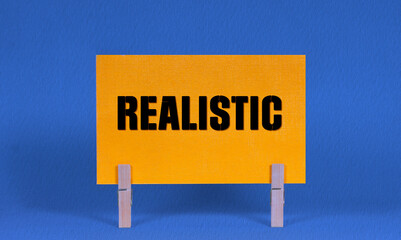 REALISTIC Symbol. Concept words REALISTIC on Yellow Rectangle Paper Held by Wooden Clothespin Blue...