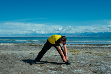 Young fitness man stretching on lake shore on summer day