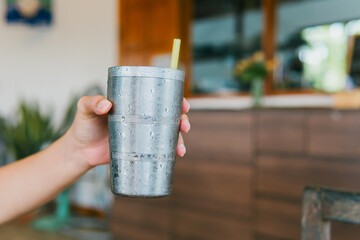 Refreshing Drink in a Metal Cup with Straw Held by Hand in a Cozy Indoor Setting