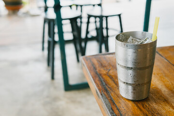 Fototapeta premium Refreshing Metallic Beverage Cup with Ice on Wooden Table in Café Environment Captured in Natural Light