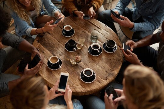 Diverse group of young adults seated around a circular wooden table, engrossed in their smartphones while enjoying coffee. Overhead shot