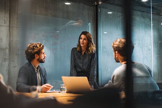 A woman leads a business meeting in a modern, dimly lit glass-walled office.  Two men listen attentively as she speaks, a laptop and notes on the table