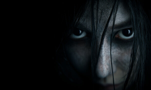 close-up of evil black eyes scary young woman with black hairs falling on her pale face; concept of horror, halloween, fear, paranormal event / promotion; black background; copy space
