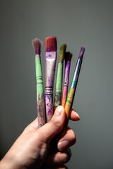 close-up of a young female hand holding used colorful paintbrushes in greens, purples, and pinks against a gray interior in natural lighting
