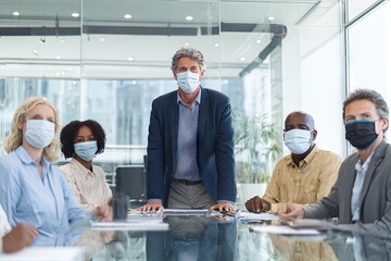 A diverse group of masked business professionals sits around a glass conference table, with a man standing prominently in the center