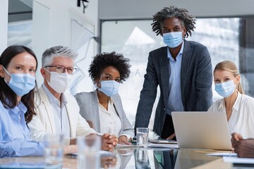 A diverse group of professionals in face masks sits around a glass conference table with a laptop, appearing confident and ready for a meeting