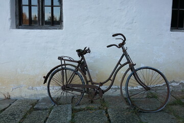 Rusty vintage bicycle leaning against an old white wall with cracked paint. The weathered setting evokes nostalgia, decay and timeless rural charm.