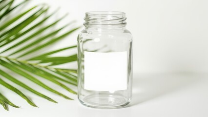 A clear glass jar with a white label sits on a white background. A green palm leaf is positioned to the left, adding a fresh and natural element to the composition’s clean, minimalistic aesthetic.