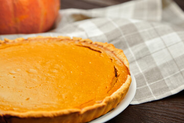 Freshly baked pumpkin pie on a white plate, placed on a wooden table with a checkered cloth, showcasing a warm and inviting autumn dessert atmosphere