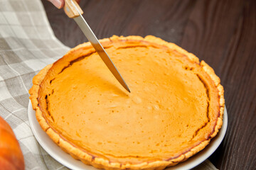 Freshly baked pumpkin pie on a white plate, with a hand holding a knife poised to slice, surrounded...
