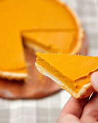 Slice of pumpkin pie held by hand, showcasing creamy filling and flaky crust, with pie on wooden...