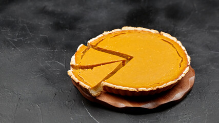 Pumpkin pie with a slice removed, showcasing a golden filling and flaky crust, placed on a wooden plate against a dark textured background, perfect for autumn desserts