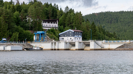Hydroelectric dam structure with utility buildings, forested hillside, and calm water. Technical...