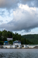 View of a dam, industrial building, and forested hillside under a large cloud-filled sky. Calm...