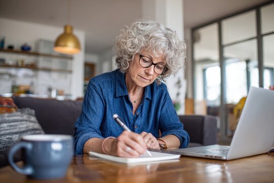 Senior woman with gray hair, glasses, and blue shirt sits at a wooden table, pensively writing in a notebook beside a laptop, a coffee mug nearby