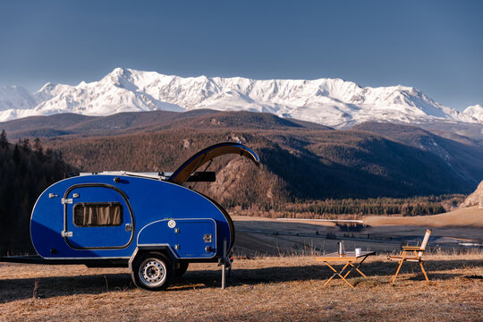 Blue trailer camper in scenic mountain landscape with chairs and snow-covered peaks. Concept vanlife in freedom trip - Powered by Adobe