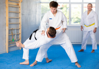 Two men practice grabbing and throwing sports mats during judo training under the guidance of an...