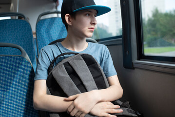 A 12 year old boy in a cap, with a backpack, sits by the window on a bus. A teenager rides alone on...