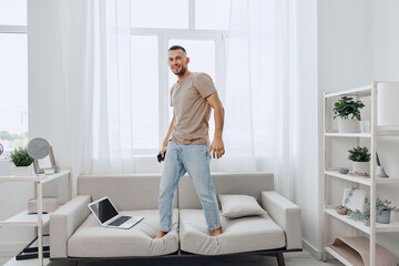 A smiling man stands on a couch, embodying the blend of work and life in a modern home. His relaxed...