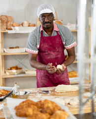 Professional male baker stands at his work bench, kneading and shaping dough to make bread,...