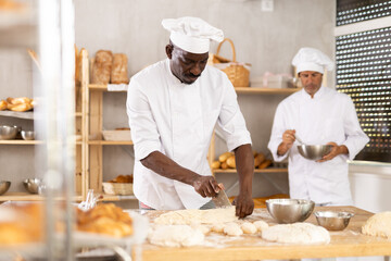 African American man works in bakery as baker, cuts dough into portions, forms pieces of dough to...