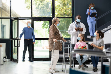 Girl visiting senior grandmother using walking frame at hospital lobby, offering flowers with love...
