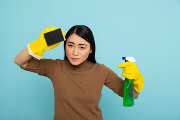 Asian housemaid with focused expression holds spray bottle and sponge, ready to tackle household...