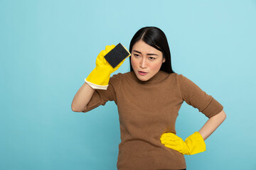 Portrait of serious asian woman in yellow rubber gloves, holding cleaning sponge and hand on waist,...
