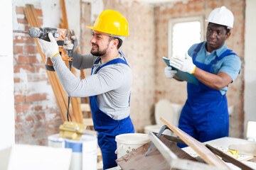 Professional builder drills a wall with a hammer drill, and his colleague makes notes, working on a...