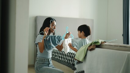 Young mother and her son are choosing clothes in bedroom, holding hangers and having fun