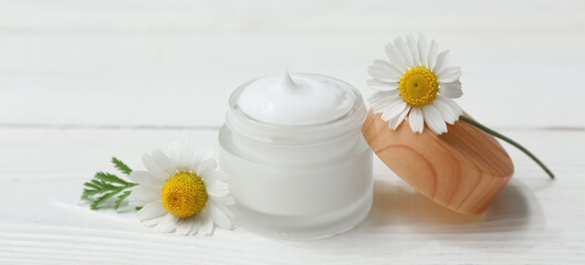 Jar of chamomile cosmetic cream and flowers on white wooden background