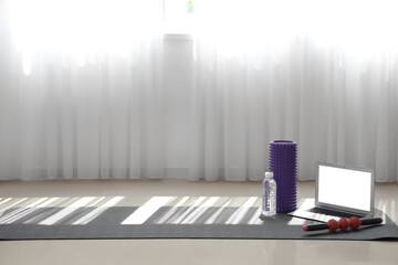 Laptop with bottle of water and rollers on fitness mat in gym