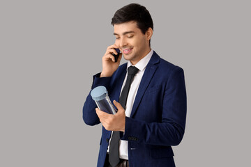 Young businessman with bottle of water talking by mobile phone on grey background