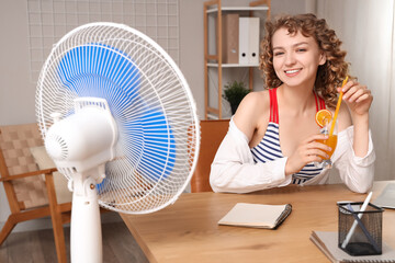 Beautiful happy young woman with electric fan and cocktail sitting on chair at workplace