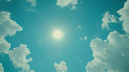 Bright sun shining through puffy white clouds in a teal sky.