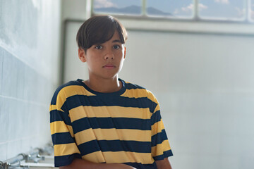 A teenage boy stands quietly indoors with a serious expression, wearing a striped shirt. Natural light comes through the window, adding a soft atmosphere.