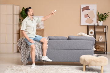 Young man switching on air conditioner while sitting on back of sofa near beige wall in living room