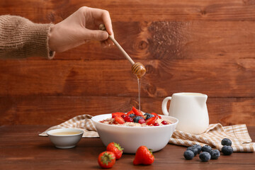 Young woman adding honey in tasty oatmeal with berries on wooden background