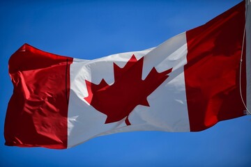 The Canadian flag is flying against the blue sky on a metal flagpole. The flag of Canada has red on its ends and white in the center, with a large red maple leaf in the middle on a white background. 