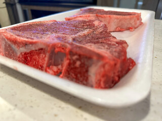 A long white styrofoam tray with two raw tender t-bone steaks. The thick grilling meat has marbling...