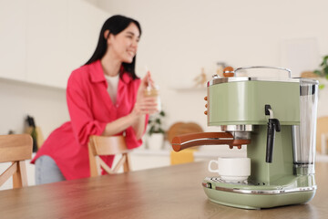 Coffee machine on table against woman with iced latte in kitchen, closeup