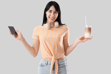 Young woman with glass of iced latte and mobile phone on light background