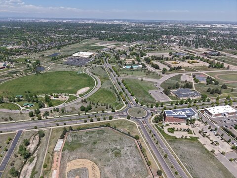 Expansive aerial drone view of Lowry, Colorado, showcasing a vibrant suburban landscape with a prominent roundabout, sports fields, green parks, commercial areas, and residential neighborhoods streets