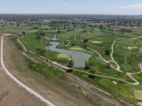 Aerial drone view of a vibrant golf course in Aurora, Colorado, featuring a large lake, meticulously manicured greens, and winding fairways. Sunny summer landscape showcases an inviting recreation