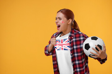 Excited fan with flags of United Kingdom on her cheek and t-shirt holding soccer ball against orange background. Space for text