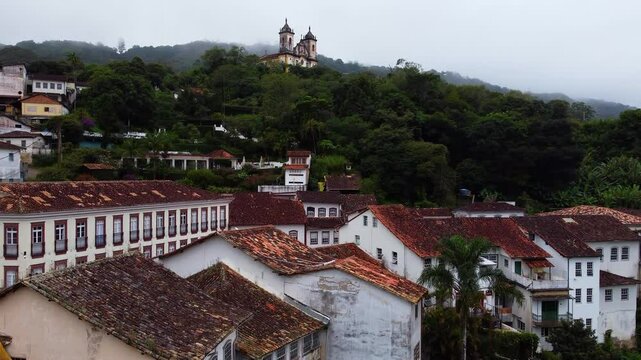 Aerial Drone Shot Over Colonial Rooftops in Ouro Preto, Brazil, Revealing Igreja Matriz de Nossa Senhora da Concei&ccedil;&atilde;o de Ant&ocirc;nio Dias on Cloudy Day