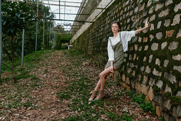 Young woman in olive green dress and white blouse posing against textured stone wall in nature,...
