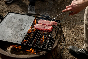 Man grilling beef steaks on a cast iron grill plate on a camp fire. Campfire cooking. Outdoor BBQ